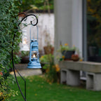 Feuerhand Baby Special 276 Sturmlaterne in blau, hängen im Garten vor grünen Pflanzen.