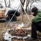 Dreibein mit Kette & Haken über einem offenen Feuer mit Kochutensilien und einem Mann, der kocht.