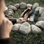 Person using Feuerrohr to tend to a campfire surrounded by stones in nature.