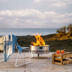 Feuerschale Tyropit in einem Garten mit lebendigem Feuer, umgeben von einem Stuhl und Getränken am Wasser.