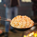 Popcornmaker mit frischem, knusprigem Popcorn über offenem Feuer, ideal für Lagerfeuerabende.